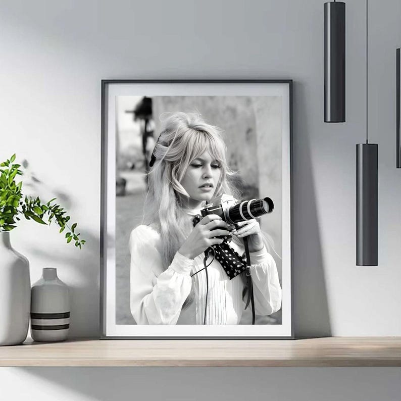 May include: Framed black and white photograph of a woman with blonde hair holding a vintage camera. She wears a white blouse. The photograph is displayed on a wooden shelf, with a vase and greenery to the left and hanging black lights to the right.