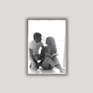 May include: Black and white framed photograph of a man and woman. The woman is seated, holding a book, while the man kneels beside her. Both are looking at each other. The photograph is set against a bright background.