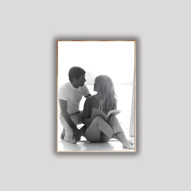 May include: Black and white framed photograph of a man and woman. The woman is seated, holding a book, while the man kneels beside her. Both are looking at each other. The photograph is set against a bright background.