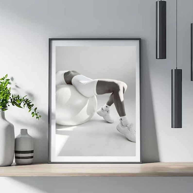May include: Black and white framed print of a person in white athletic wear, leaning back on a white exercise ball. The person is wearing white sneakers and socks. The image is displayed on a wooden shelf with decorative vases and hanging black light fixtures.