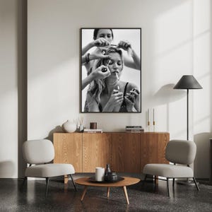 May include: Black and white framed print of a woman smoking a cigarette while having her hair styled. The image is displayed above a wooden cabinet, with two gray chairs and a round coffee table in front.