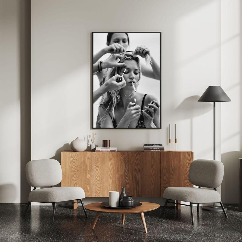 May include: Black and white framed print of a woman smoking a cigarette while having her hair styled. The image is displayed above a wooden cabinet, with two gray chairs and a round coffee table in front.