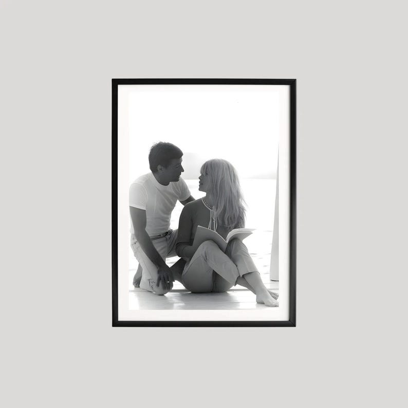 May include: Black and white framed photograph of a man and woman. The man is kneeling, looking at the woman, who is seated and holding a book. The photograph is in a black frame.