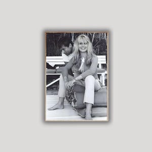 May include: Black and white framed photograph of a woman and a man sitting on a bench. The woman is wearing a long-sleeved top and pants. The man is wearing a white t-shirt and pants. The photograph is in a wooden frame.
