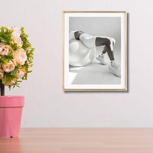 May include: A framed black and white photograph of a person in white athletic wear, leaning back on a white exercise ball. The person is wearing white sneakers and socks. The frame is gold.