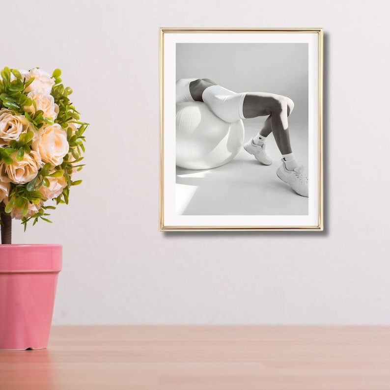 May include: A framed black and white photograph of a person in white athletic wear, leaning back on a white exercise ball. The person is wearing white sneakers and socks. The frame is gold.