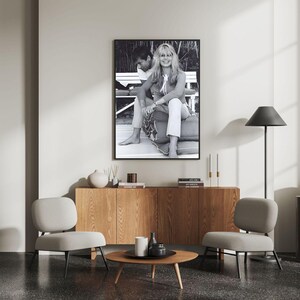 May include: A framed black and white photograph of a couple sitting on a bench, displayed above a wooden cabinet. The room features two gray chairs, a round coffee table, and a black floor lamp. Decorative vases and candles are also present.