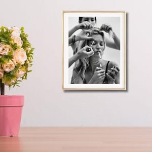 May include: Black and white framed photograph of a person being styled. Several hands are visible, applying makeup and styling hair. The person is smoking a cigarette. The photograph is in a gold frame and is hanging on a white wall.