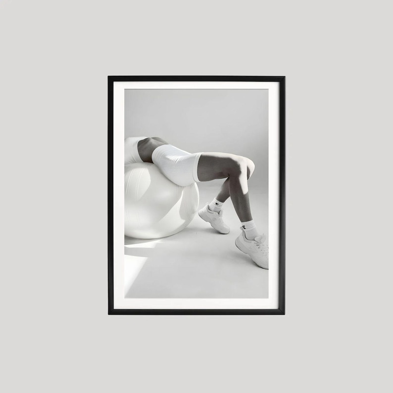 May include: Black and white framed print featuring a person in white athletic wear, resting on a white exercise ball. The image is a minimalist composition, emphasizing form and light, suitable for fitness or wellness themes.