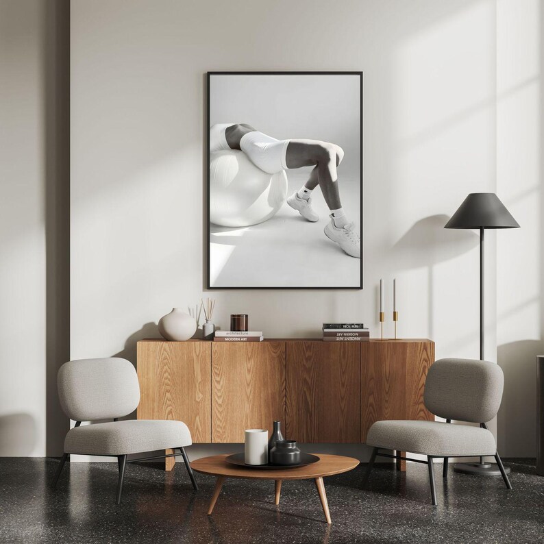 May include: A framed black and white photograph of a person in athletic wear on an exercise ball. The photograph hangs above a wooden cabinet with decorative items. Two gray chairs and a round coffee table complete the room's decor.