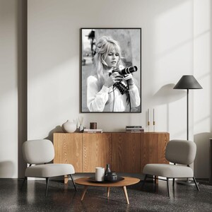 May include: Monochrome framed print featuring a woman with blonde hair holding a camera. She wears a white blouse. The print hangs above a wooden cabinet, with two grey chairs and a round coffee table in the room.