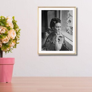 Pu&ograve; includere: Fotografia in bianco e nero incorniciata di una persona che fuma una sigaretta, seduta vicino a una finestra. La persona indossa un vestito a scacchi e ha i capelli acconciati con un fiore. La cornice &egrave; dorata e l'immagine &egrave; esposta su una parete bianca.