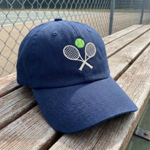 May include: Navy blue baseball cap with a curved brim. The cap features an embroidered design of two crossed tennis rackets and a green tennis ball. The hat is sitting on a wooden bench.