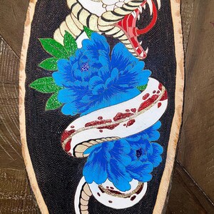 May include: A white snake with red and gold accents is coiled around two blue flowers on a black background. The snake's mouth is open and its fangs are visible. The artwork is painted on a wooden slice.