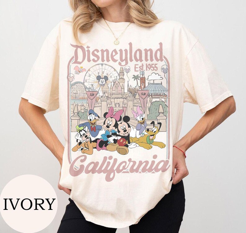 May include: An ivory short-sleeve t-shirt featuring a graphic print of Disneyland California Est. 1955. The design includes illustrations of Mickey Mouse, Minnie Mouse, Donald Duck, Daisy Duck, Goofy, and Pluto in front of the castle and ferris wheel.