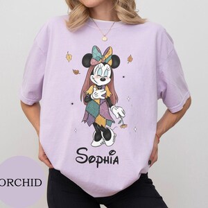 May include: A light purple t-shirt featuring a graphic of a cartoon mouse character dressed in a patchwork-style dress and bow, with long hair and stitched facial details. The name Sophia is printed in black script below the character illustration.