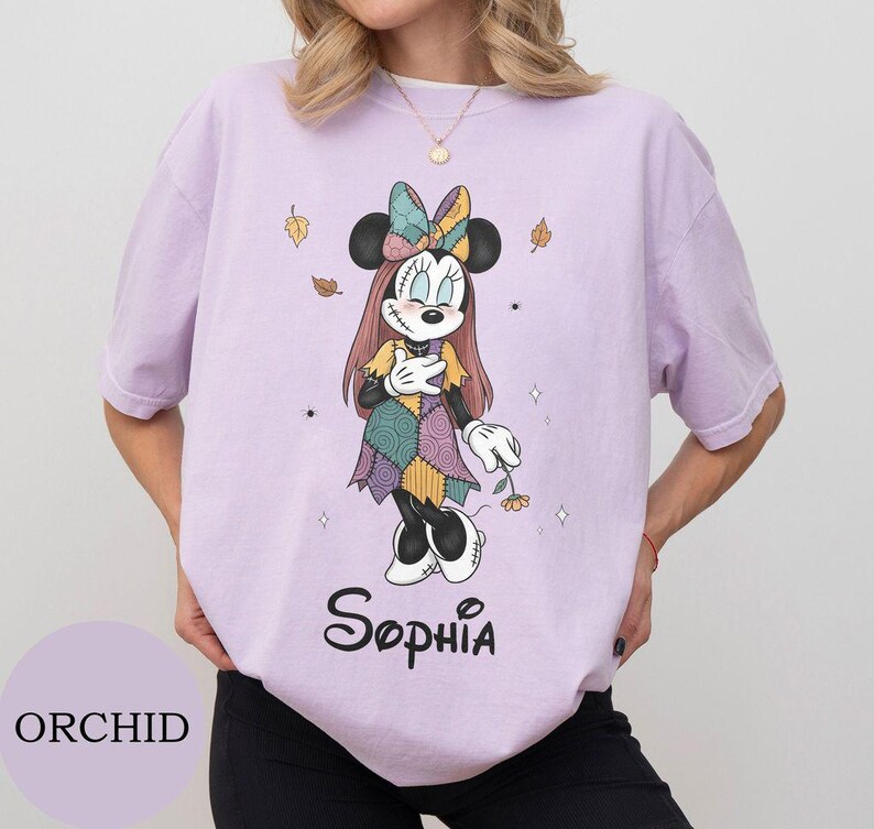 May include: A light purple t-shirt featuring a graphic of a cartoon mouse character dressed in a patchwork-style dress and bow, with long hair and stitched facial details. The name Sophia is printed in black script below the character illustration.