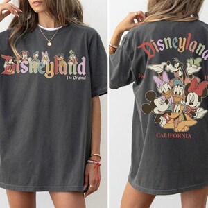 Puede incluir: Camiseta gris oscuro con el logotipo de Disneyland en letras coloridas y personajes de Disney. La parte trasera de la camiseta muestra el logotipo de Disneyland y varios personajes de Disney, con "California" y "1955" impresos debajo.