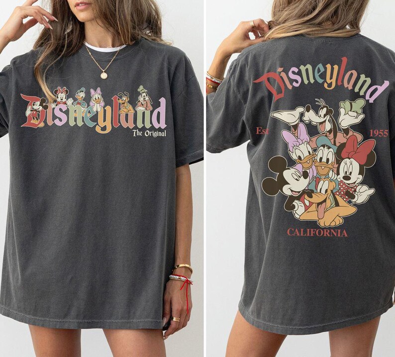 Puede incluir: Camiseta gris oscuro con el logotipo de Disneyland en letras coloridas y personajes de Disney. La parte trasera de la camiseta muestra el logotipo de Disneyland y varios personajes de Disney, con "California" y "1955" impresos debajo.