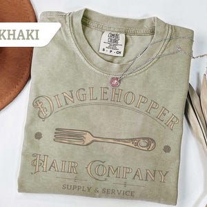 May include: Khaki-coloured t-shirt with the text "Dinglehopper Hair Company" and a fork graphic. The shirt has a crew neck and short sleeves. A silver necklace with a pendant is on the shirt.