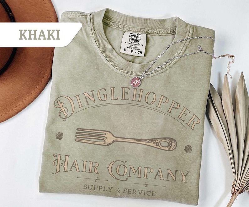 May include: Khaki-coloured t-shirt with the text "Dinglehopper Hair Company" and a fork graphic. The shirt has a crew neck and short sleeves. A silver necklace with a pendant is on the shirt.