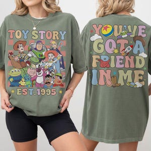 May include: Olive green t-shirt featuring a colorful graphic of Toy Story characters, the text "TOY STORY EST. 1995" on the front, and "YOU'VE GOT A FRIEND IN ME" on the back. The shirt has a relaxed fit.