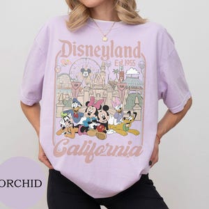 May include: A light purple t-shirt featuring a graphic print of Disneyland California, established in 1955. The design includes Mickey Mouse, Minnie Mouse, Donald Duck, Daisy Duck, Goofy, and Pluto in front of the castle and a Ferris wheel.