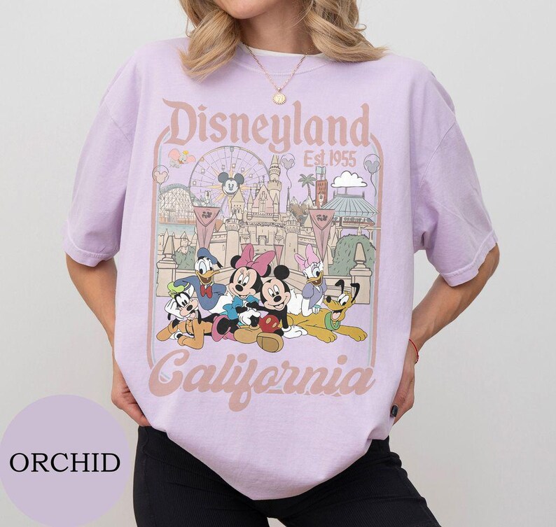 May include: A light purple t-shirt featuring a graphic print of Disneyland California, established in 1955. The design includes Mickey Mouse, Minnie Mouse, Donald Duck, Daisy Duck, Goofy, and Pluto in front of the castle and a Ferris wheel.