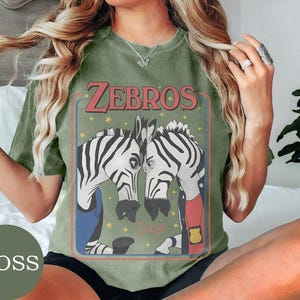 May include: A moss green crew neck t-shirt featuring a graphic print of two zebras facing each other. The text "ZEBROS" is printed in red above the zebras, which are depicted wearing clothing and a badge. Small yellow stars surround the zebras.