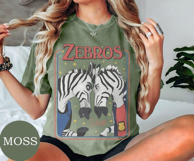 May include: A moss green crew neck t-shirt featuring a graphic print of two zebras facing each other. The text "ZEBROS" is printed in red above the zebras, which are depicted wearing clothing and a badge. Small yellow stars surround the zebras.