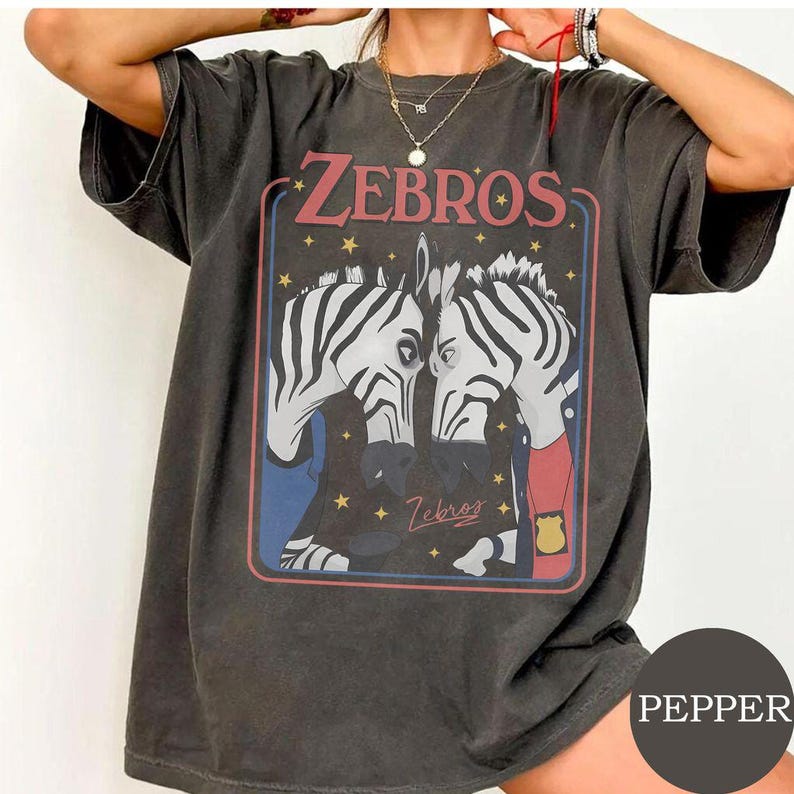 May include: A charcoal gray short-sleeve t-shirt featuring a graphic print of two zebras facing each other. The text "ZEBROS" appears at the top in red, with small yellow stars scattered around the zebras. One zebra wears a red shirt and a gold badge.