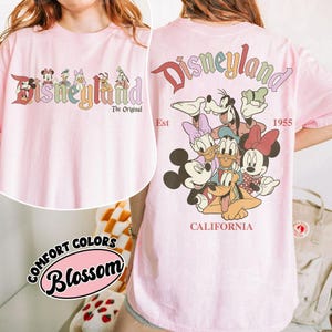 Puede incluir: Camiseta rosa claro con la palabra "Disneyland" en una fuente retro colorida y personajes de Disney. La parte trasera de la camiseta tiene el logo de Disneyland, los personajes y el texto "California 1955".