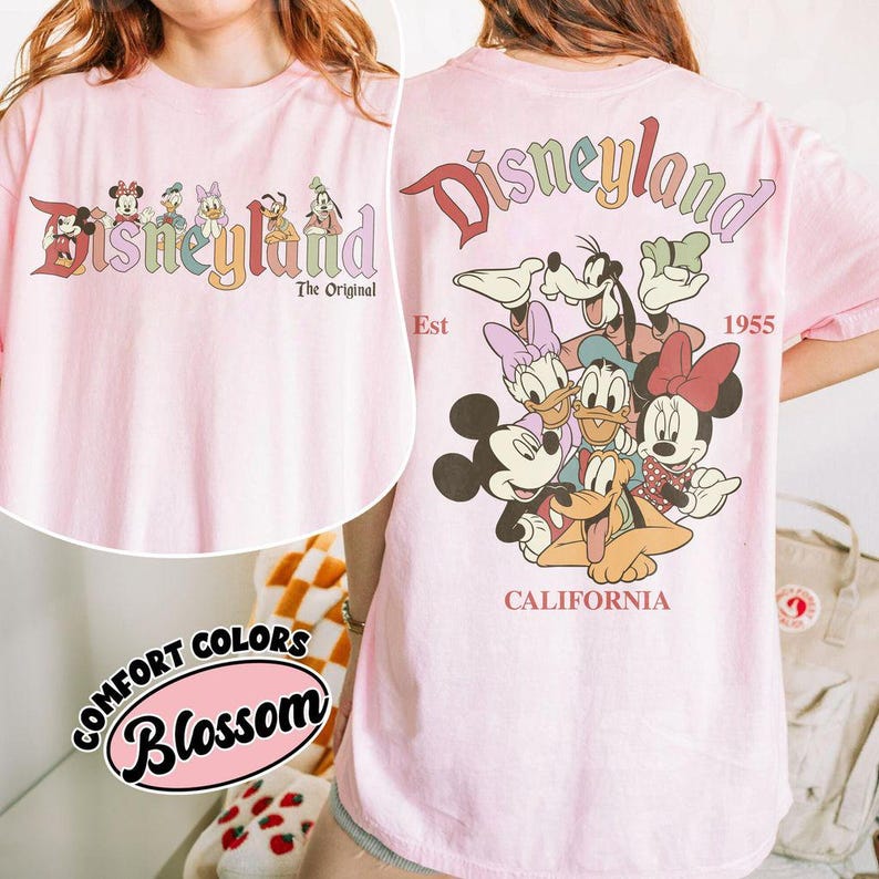 Puede incluir: Camiseta rosa claro con la palabra "Disneyland" en una fuente retro colorida y personajes de Disney. La parte trasera de la camiseta tiene el logo de Disneyland, los personajes y el texto "California 1955".