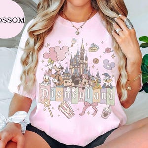May include: A light pink short-sleeve t-shirt featuring a graphic print of a castle, Mickey Mouse, Minnie Mouse, Goofy, Donald Duck, and Daisy Duck. The text Disneyland is printed in a stylized font above icons of popcorn, a bow, and a drink.