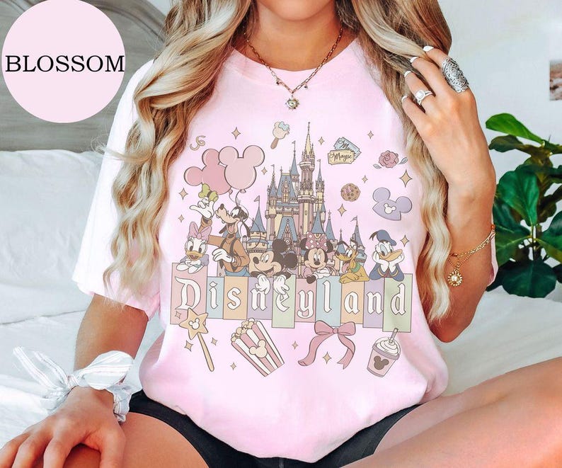 May include: A light pink short-sleeve t-shirt featuring a graphic print of a castle, Mickey Mouse, Minnie Mouse, Goofy, Donald Duck, and Daisy Duck. The text Disneyland is printed in a stylized font above icons of popcorn, a bow, and a drink.