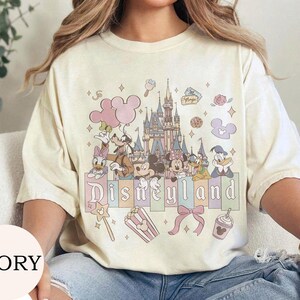 May include: An ivory short-sleeve t-shirt featuring a graphic print of a castle, Mickey Mouse, Minnie Mouse, Goofy, Donald Duck, and Daisy Duck. The design includes pastel-colored balloons, a bow, popcorn, and the text Disneyland in a serif font.