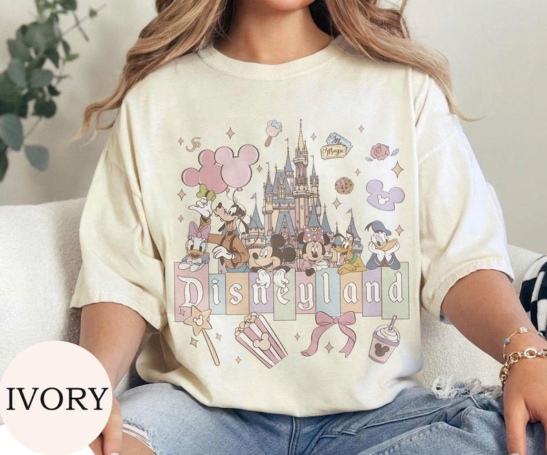 May include: An ivory short-sleeve t-shirt featuring a graphic print of a castle, Mickey Mouse, Minnie Mouse, Goofy, Donald Duck, and Daisy Duck. The design includes pastel-colored balloons, a bow, popcorn, and the text Disneyland in a serif font.