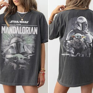 May include: A charcoal gray short-sleeve t-shirt featuring a graphic print of The Mandalorian and Grogu. The front displays the Star Wars The Mandalorian logo above the characters, while the back shows a larger illustration of the same duo.