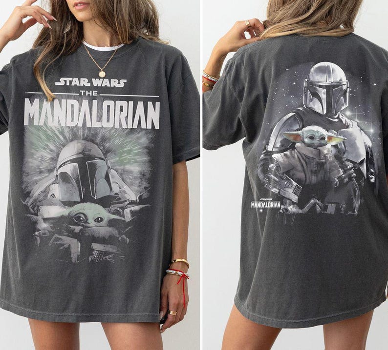 May include: A charcoal gray short-sleeve t-shirt featuring a graphic print of The Mandalorian and Grogu. The front displays the Star Wars The Mandalorian logo above the characters, while the back shows a larger illustration of the same duo.