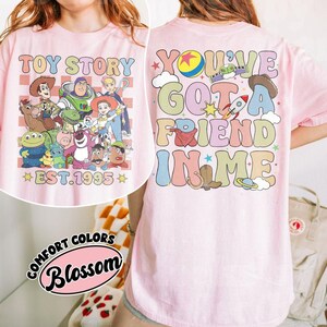 May include: Pink t-shirt featuring a colorful graphic of Toy Story characters and the text "TOY STORY EST. 1995" on the front. The back of the shirt displays "YOU'VE GOT A FRIEND IN ME" with related imagery.