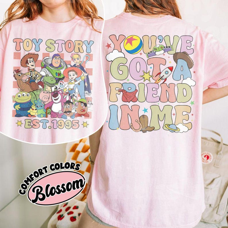 May include: Pink t-shirt featuring a colorful graphic of Toy Story characters and the text "TOY STORY EST. 1995" on the front. The back of the shirt displays "YOU'VE GOT A FRIEND IN ME" with related imagery.
