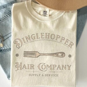 May include: Cream-coloured t-shirt with the text "Dinglehopper Hair Company" in a vintage font, featuring a fork graphic. The shirt also includes the words "Supply & Service". A Comfort Colors tag is visible at the neckline.