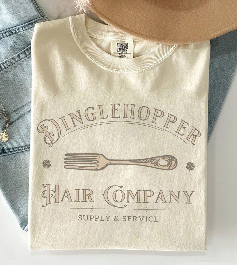 May include: Cream-coloured t-shirt with the text "Dinglehopper Hair Company" in a vintage font, featuring a fork graphic. The shirt also includes the words "Supply & Service". A Comfort Colors tag is visible at the neckline.