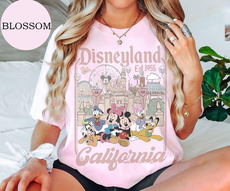 May include: A light pink short-sleeve t-shirt featuring a graphic print of Disneyland California Est. 1955. The design includes illustrations of Mickey Mouse, Minnie Mouse, Donald Duck, Daisy Duck, Goofy, and Pluto in front of the castle.