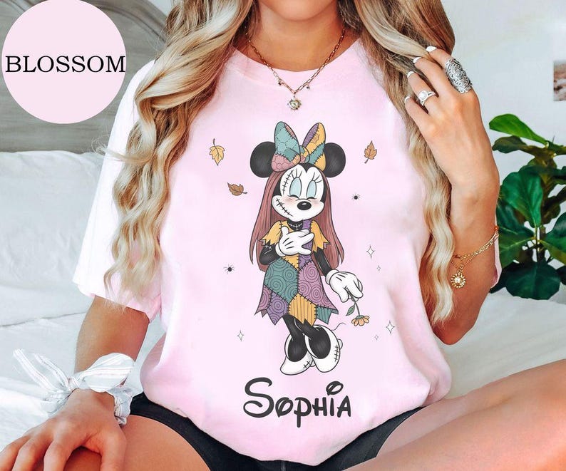 May include: A light pink t-shirt featuring a graphic of a cartoon mouse character dressed in a patchwork dress and bow, with stitched facial details, holding a small flower. The name Sophia is printed in black script below the character graphic.