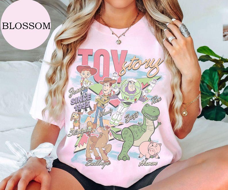 May include: A light pink short-sleeve t-shirt featuring a graphic print of Toy Story characters including Woody, Buzz Lightyear, Jessie, Rex, Slinky Dog, Bullseye, and Hamm, with the text Toy Story, Jessie, Since 1995, Woody, Buzz, Rex, Slinky Dog, Bullseye, and Hamm.