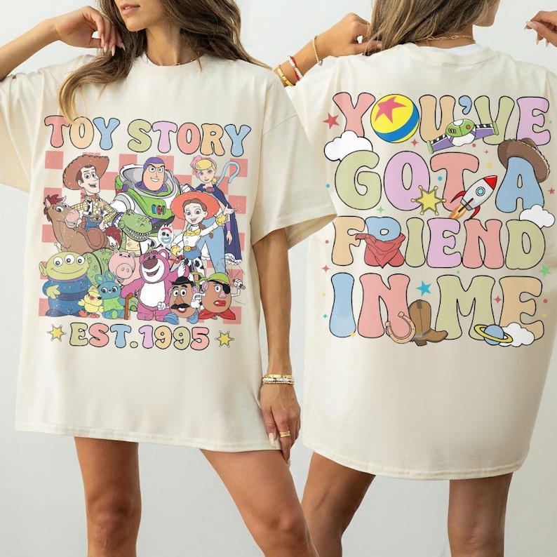 May include: Cream-colored t-shirt featuring a colorful graphic of Toy Story characters and the text "TOY STORY EST. 1995" on the front. The back of the shirt displays the phrase "YOU'VE GOT A FRIEND IN ME" with related illustrations.