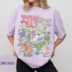 May include: A light purple t-shirt featuring a graphic print of Toy Story characters including Jessie, Woody, Buzz Lightyear, Rex, Slinky Dog, Bullseye, and Hamm. The text reads Toy Story, Jessie Since 1995, Woody, Buzz, Rex, Bullseye, and Hamm.