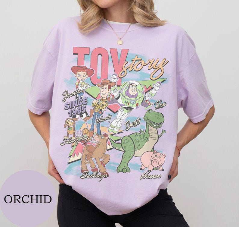 May include: A light purple t-shirt featuring a graphic print of Toy Story characters including Jessie, Woody, Buzz Lightyear, Rex, Slinky Dog, Bullseye, and Hamm. The text reads Toy Story, Jessie Since 1995, Woody, Buzz, Rex, Bullseye, and Hamm.