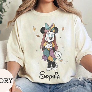 May include: An ivory t-shirt featuring a graphic of a character resembling Minnie Mouse dressed in a patchwork-style dress and bow, with long hair and stitched details. The name Sophia is printed in black script below the character illustration.